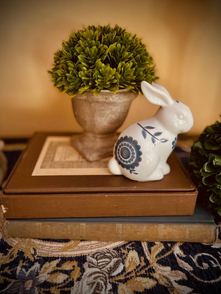 Spring Easter decor chinoiserie ceramic bunny rabbit next to potted boxwood topiary 4' faux plant decor sitting on top of stacked vintage books on table
